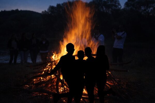 People silhouetted around a large bonfire with bright orange flames against a darkening sky, with trees and other figures visible in the background.