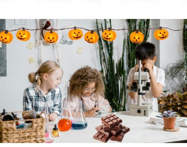 Three children conducting science experiments at a table with microscope, beakers and chocolate, beneath Halloween pumpkin string lights.