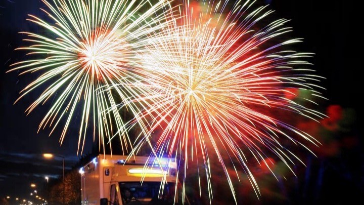 Colourful fireworks display with red, green and white bursts exploding in the night sky above buildings with illuminated windows.