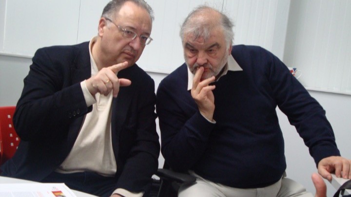 Two men in dark jackets sitting at a white table, both making shush gestures with their fingers to their lips.