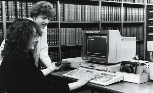 Two people using an early computer terminal in a library, surrounded by shelves of reference books.