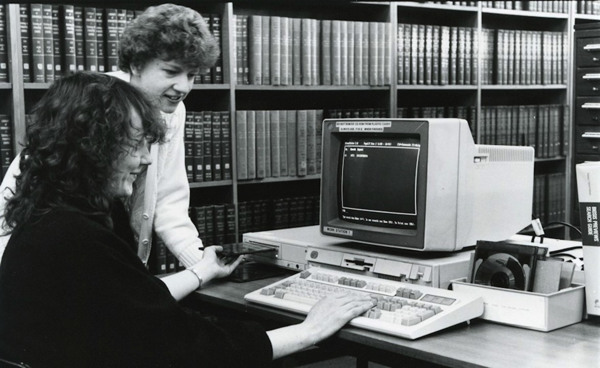 Two people using an early computer terminal in a library, surrounded by shelves of reference books.