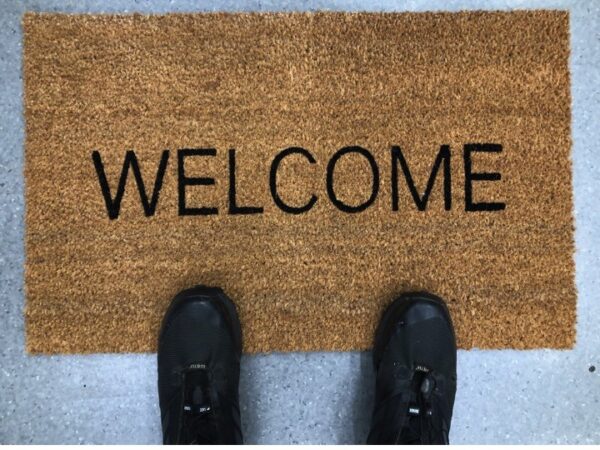 Person's feet in black shoes standing on a brown doormat with WELCOME text on a concrete floor.