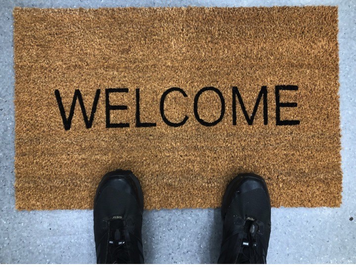 Person's feet in black shoes standing on a brown doormat with WELCOME text on a concrete floor.
