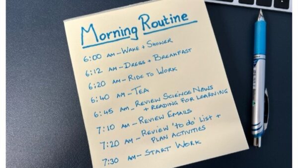 A handwritten morning routine schedule on a yellow sticky note beside a laptop and pen, listing times from 6:00 AM to 7:30 AM with daily tasks.