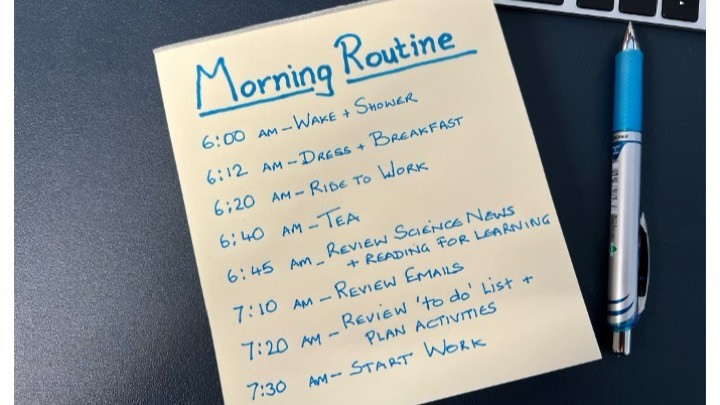 A handwritten morning routine schedule on a yellow sticky note beside a laptop and pen, listing times from 6:00 AM to 7:30 AM with daily tasks.