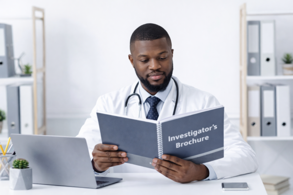 A doctor in a white coat and stethoscope reading an Investigator's Brochure at a desk with a laptop in a modern office setting.