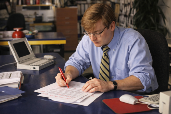 Man in blue shirt and tie editing document with red pen at desk, with laptop and papers in office setting.